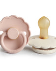 The FRIGG Daisy Natural Rubber Pacifier 2-Pack features two daisy-shaped pacifiers in soft pastels, with one upright showing a natural latex nipple and white base, and the other flat revealing a pale pink front with embossed FRIGG branding.