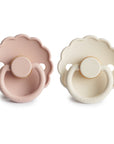 The FRIGG Daisy Natural Rubber Pacifier 2-Pack by FRIGG features two daisy-shaped pacifiers, one in muted pink and one in light cream, both with round handles, embossed fronts, and natural rubber latex nipples for gentle soothing.
