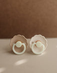 Two Frigg Daisy Night pacifiers in Cream and Croissant on a beige surface.
