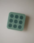 A green silicone mushie Fresh Food Feeder Freezer Tray with nine round compartments for baby food storage rests on a plain white surface. The word “mushie” is embossed in the center of the tray.
