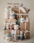 A stylish display of mushie's Deluxe Whales/Tradewinds Feeding Kit—featuring pastel cups, plates, bowls, utensils, and teethers—arranged on ribbon-wrapped gift boxes for a soft, elegant look.