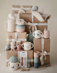 A display of neatly stacked, gift-wrapped boxes with mushie's Stage 1 Bees Feeding Kit—cups, bowls, spoons, and toys in pastel silicone featuring cute animal designs and soft tones—arranged attractively in front.