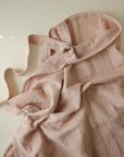 The mushie Muslin Nursing Cover in pale pink rests crumpled on a light surface, with neck and waist straps draped across the soft organic cotton, while gentle natural light highlights its texture.