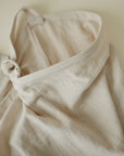 A close-up of the mushie Muslin Nursing Cover, featuring a light beige textured organic cotton bag with a knotted strap, shown against a neutral background.