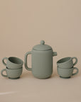 The Mushie Silicone Tea Play Set features a matte green teapot with a lid, centered between two stacks of ribbed teacups—ideal for pretend play in any play kitchen—displayed against a simple beige background.