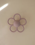 The mushie Daisy Rattle Teether, a pastel purple flower-shaped baby teething toy with six loops and the brand name embossed in the center, rests on a light beige surface.