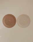 Two round mushie Silicone Drain Stoppers, featuring concentric circle designs in brown and beige, are placed side by side on a neutral surface—ideal as stylish bath accessories or functional drain covers.