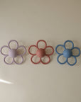 Three mushie Daisy Rattle Teethers in pastel purple, pink, and blue are arranged in a row on a neutral background. Each silicone teether features six textured petals, with soft lighting creating gentle shadows.