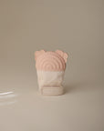 The mushie Rainbow Teething Mitten features a soft beige fabric, a light pink silicone top with a rainbow pattern, and small bear ear accents, displayed upright against a plain light background.
