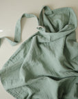 The mushie Muslin Nursing Cover in light green, with a neck strap and waist tie, lies gently wrinkled and partially folded on a pale surface, highlighting its breathable fabric.