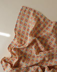 The mushie Organic Cotton Muslin Swaddle Blanket features a colorful geometric pattern of small squares and circles in orange, yellow, blue, and white on soft organic cotton, displayed on a light background with delicate folds.