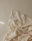 The mushie Organic Cotton Muslin Swaddle Blanket features soft, crumpled organic cotton with a subtle brown twig pattern on beige—perfect for swaddling or use as a cozy baby blanket.