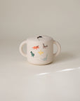 A cream-colored, spill-proof Snack Cup by mushie with two handles and colorful dinosaur illustrations sits on a plain, light background—perfect for toddlers.