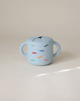 The mushie Snack Cup is a spill-proof, light blue silicone cup with two handles for toddlers, decorated with colorful car illustrations and shown on a neutral background.