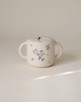 The mushie Snack Cup is a cream-colored ceramic container for toddlers, featuring two handles, a lid, and delicate blue floral designs. It’s shown on a neutral background with soft lighting.