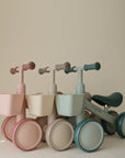 Three mushie Mini Toddler Bikes in pastel colors are lined up against a beige background.