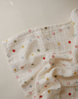 The mushie Organic Cotton Muslin Swaddle Blanket features soft white crinkled fabric with scattered colorful circles and smiley faces, perfect for gently wrapping your baby in comfort.