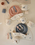 A flat lay of mushie's Deluxe Whales/Tradewinds Feeding Kit, featuring pastel pink, blue, and cream cups, bowls, divided plates, and spoons with charming animal and bird motifs—perfect toddler feeding essentials with matching utensils.