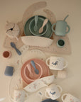A flat lay of mushie’s Deluxe Vanilla Confetti/Cambridge Blue Feeding Kit, featuring pastel plates, bowls, spoons, cups, and divided placemats with animal and floral designs arranged neatly on a beige surface.