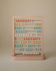 The Mushie Wooden Abacus features pastel yellow, pink, blue, and white beads on metal rods—a perfect Montessori counting toy for toddlers.