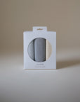 A white box with a circular window shows three folded Mushie Changing Pad Liners in gray, light blue, and cream. The Mushie brand name is printed on the box against a plain, neutral background.