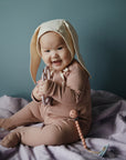 A happy baby in mushie Zipper Pajama and a mauve bunny ear hat sits on a purple blanket, holding a teething ring toy with both hands against a muted blue background.