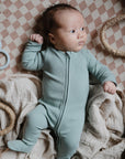 A baby in mushie Zipper Pajama rests on a beige checkered blanket in a woven basket, with a soft cloth and blue bunny-shaped teether nearby.