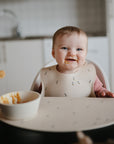 A smiling baby sits in a high chair, wearing a mushie Silicone Baby Bib. Food is on their face, with a bowl and spoon on the tray. Kitchen sink and cabinets are visible in the background.