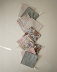 Seven mushie Muslin Cotton Washcloths in assorted pastel floral and solid patterns are arranged diagonally on a light beige surface, each with a small hanging loop.