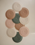Twelve mushie Silicone Drain Stoppers in beige, cream, and green are arranged in a rectangle. Each has concentric raised circles, inspired by bath drain covers, for a textured and modern look.