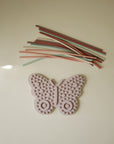 Mushie Silicone Sensory Toy, pink butterfly, under pastel silicone strings on beige surface.