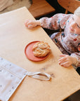 A young child in a patterned smock sits at a wooden table with a cinnamon pastry on an orange plate and a mushie Water Resistant Wet Bag for diaper bag storage nearby.