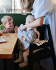 In a bright kitchen, a woman in a blue dress helps a baby wearing the mushie Long Sleeve Bib in a high chair, while a smiling toddler sits at a nearby wooden table.