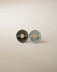 The FRIGG Little Viking Silicone 2-Pack by FRIGG includes two round, medical-grade silicone pacifiers—one dark green and one light blue—standing upright on a softly lit beige surface against a neutral background. Made in Denmark.
