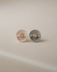 The FRIGG Little Viking Silicone 2-Pack by FRIGG features two round pacifiers—one beige, one light gray—standing upright side by side on a softly lit neutral background.
