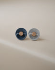 The FRIGG Little Viking Silicone 2-Pack by FRIGG includes two round pacifiers—one dark blue, one light gray—with beige nipples. Made in Denmark from medical-grade silicone, they are displayed side by side on a neutral surface under soft lighting.