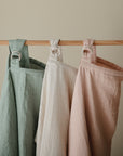 Three mushie Muslin Nursing Covers in sage green, off-white, and blush pink hang from a wooden rod by metal rings against a beige background.