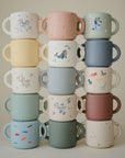 A set of colorful, two-handled mugs with animal and nature designs is arranged in three columns beside a mushie Snack Cup—a spill-proof silicone cup—against a light background, ideal for toddlers.
