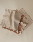 Four mushie Muslin Cotton Washcloths in soft neutrals are shown on a plain backdrop; one displays embroidered rainbows, two include edge fabric loops, and all feature subtle textures.