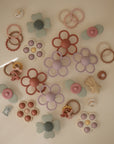 The mushie Daisy Rattle Teether, along with other pastel silicone flower and ring teethers, is displayed in a flat lay on a light surface in soft green, pink, beige, and purple hues.
