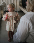 A small child in a cream dress with red hearts holds a pink mushie Silicone Baby Food Container and a cup, standing barefoot indoors. A blonde-haired adult sits nearby, slightly out of focus in the foreground.