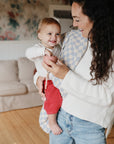 A smiling woman with long curly hair holds a happy baby in a cozy living room. Nearby, mushie Silicone Baby Food Containers made from food-grade silicone sit on a table, with a beige couch and floral wallpaper in the background.