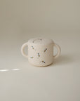 The mushie Snack Cup, a cream-colored ceramic cup with two handles designed for toddlers and adorned with small painted bees, is displayed on a plain, softly lit surface with a minimal background.