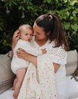 A woman in a white dress sits on a cushioned outdoor bench, holding a baby wrapped in a mushie Organic Cotton Muslin Swaddle Blanket. Surrounded by lush greenery, they smile warmly, the colorful blanket brightening the scene.
