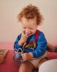 A smiling toddler with curly hair sits on a pink surface, holding the mushie Daisy Rattle Teether. Wearing a blue rainbow sweater, the child is near an abacus and an adult’s tattooed arm.