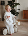 Smiling toddler rides a beige mushie Mini Toddler Bike indoors near plants and furniture.