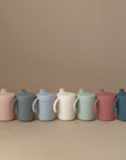 Seven Mushie Trainer Sippy Cups in pastel pink, gray, blue, cream, white, mint green, and mauve are lined up on a beige background. Each non-toxic cup has easy-grip handles and a spout lid for little hands.