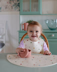 A smiling toddler wears a mushie Silicone Baby Bib with an adjustable neck strap, sitting at a table with a silicone cup in a cozy kitchen with pastel green cabinets and floral wallpaper.