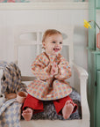 A smiling toddler in a patterned shirt and red pants wears a mushie Long Sleeve Bib while sitting on a white chair with a gray cushion, surrounded by toy cups and a flower vase on a green shelf in a cozy room.