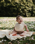 A smiling baby wears a mushie Silicone Baby Bib while sitting on a blanket outdoors, surrounded by grass and white petals, with trees in the sunny background.